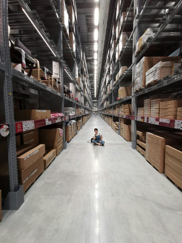 Transfer Printing for Stock Management: The Guide to Turning Dead Stock into Cash An Asian woman sits in a large warehouse aisle filled with inventory racks and shelves.